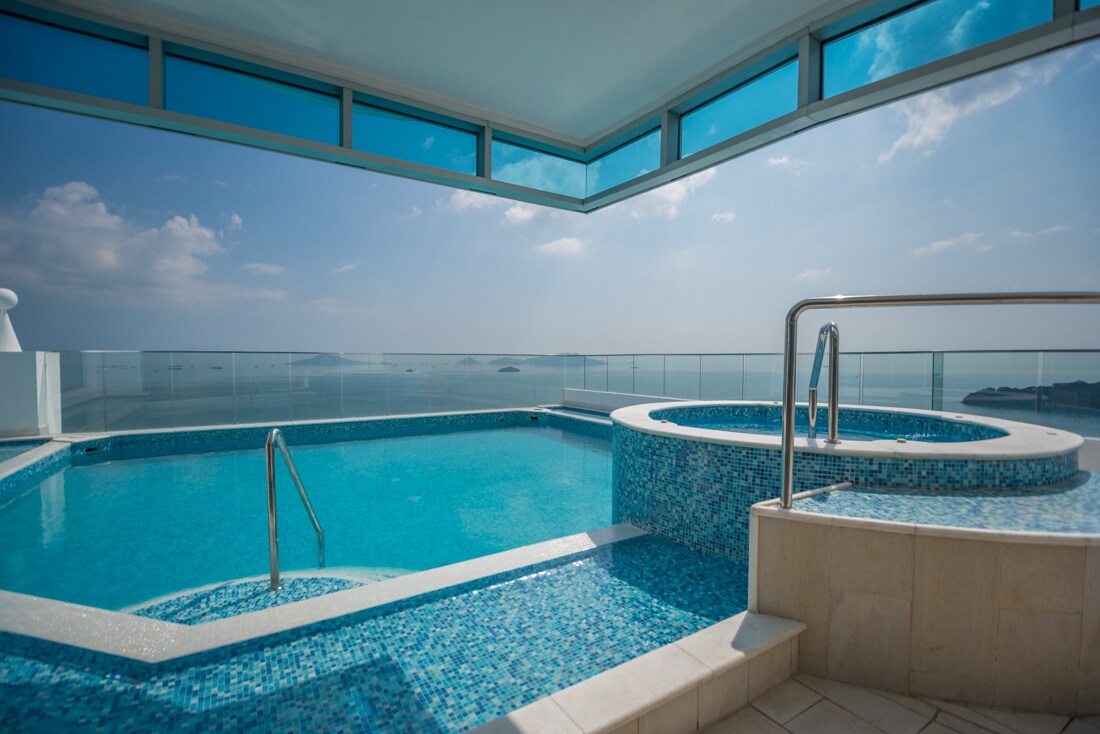 The rooftop pool area features a large swimming pool alongside a circular jacuzzi, both framed by expansive glass walls. Bright turquoise water reflects the sunlight, while panoramic views of the ocean and distant islands provide a stunning backdrop.