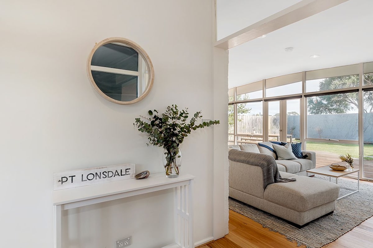 A welcoming entryway is complemented by a round mirror and a small console table displaying a vase of greenery. The open living area features large windows that offer views of the deck and grassed area, enhancing the sense of spaciousness.