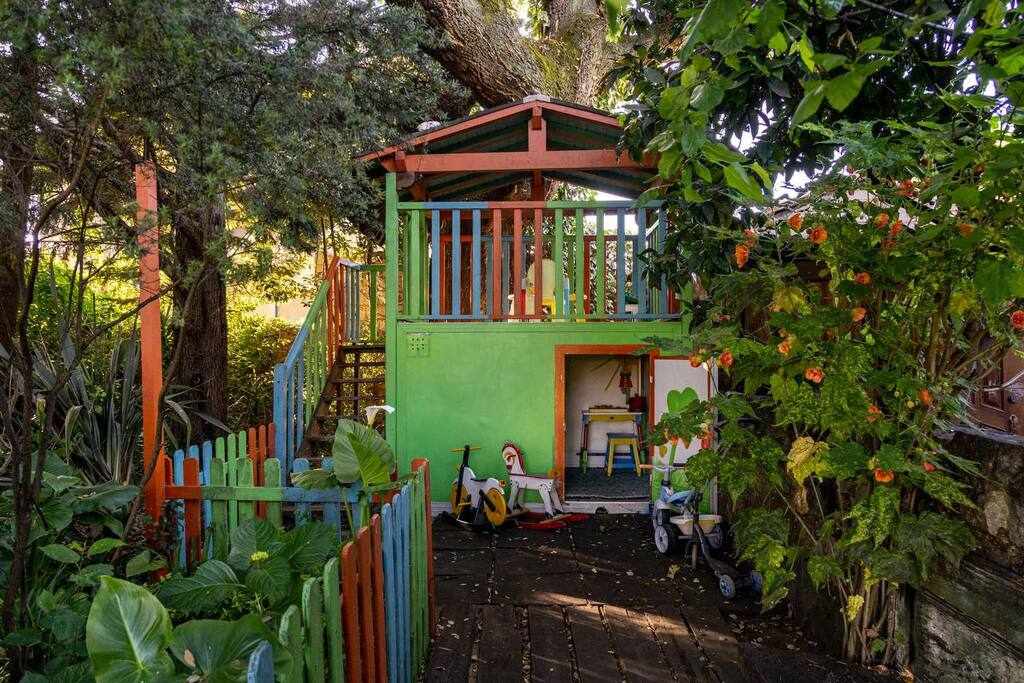 A cheerful children’s playhouse is nestled among vibrant greenery, featuring a colorful exterior with a green and orange roof. A set of steps leads up to a welcoming entrance, while various toys, including a tricycle and a doll, can be seen in the play area.