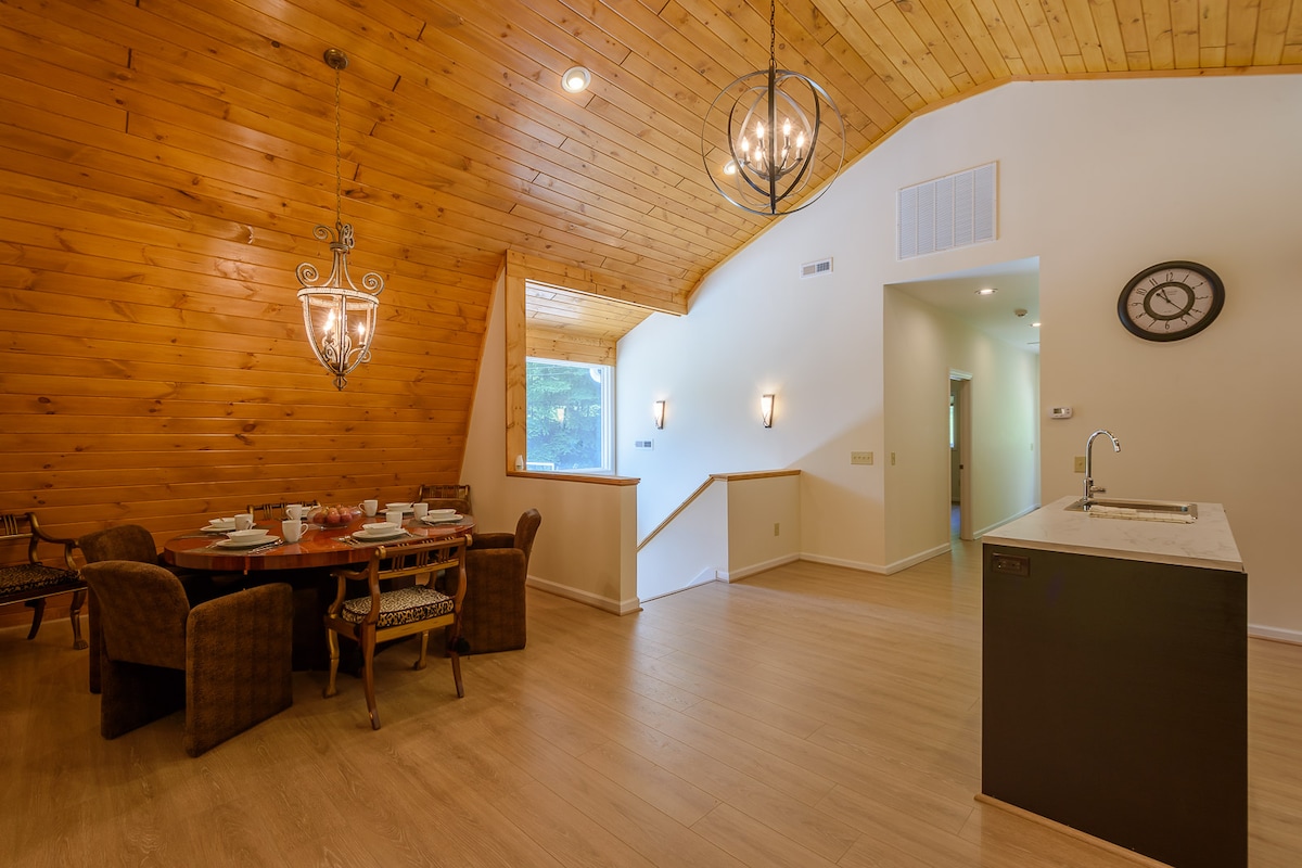 An inviting dining area features a round wooden table set for six, with plush chairs surrounding it. A high wooden ceiling with decorative lighting adds warmth to the space. The open layout leads to a modern kitchen area, with a simple sink and a wall clock visible.