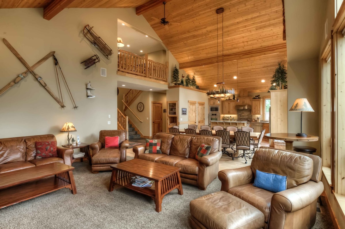 An open great room features high vaulted ceilings and warm wooden beams. Leather furniture is arranged around a central coffee table, while decorative sleds adorn the walls. The kitchen and dining area are visible in the background, creating a spacious and inviting gathering space.