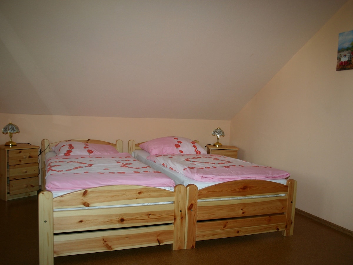 The room features two wooden beds with light pink bedding decorated with floral patterns. Bedside tables with lamps are positioned next to each bed. Soft natural light filters in, highlighting the warm wooden tones of the furniture against the light-colored walls.