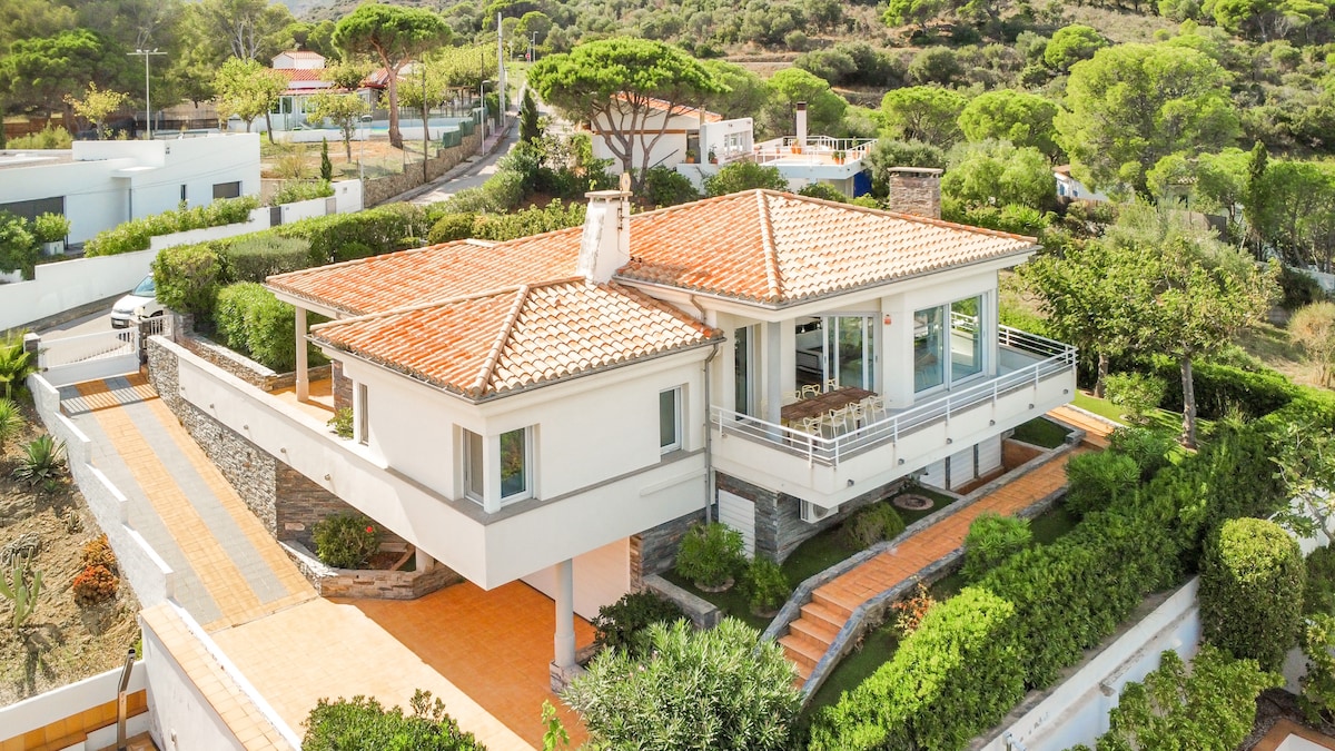 A spacious villa is shown, featuring a charming terracotta-tiled roof and large windows. The property is surrounded by lush greenery and manicured gardens, creating a serene environment. A patio area with outdoor seating is visible, providing a perfect space for relaxation and enjoying the views.