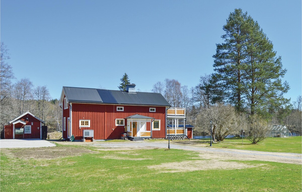 A red two-story vacation home is set against a clear blue sky, surrounded by a spacious green lawn. A smaller outbuilding is visible nearby, bordered by trees, indicating a peaceful outdoor environment.