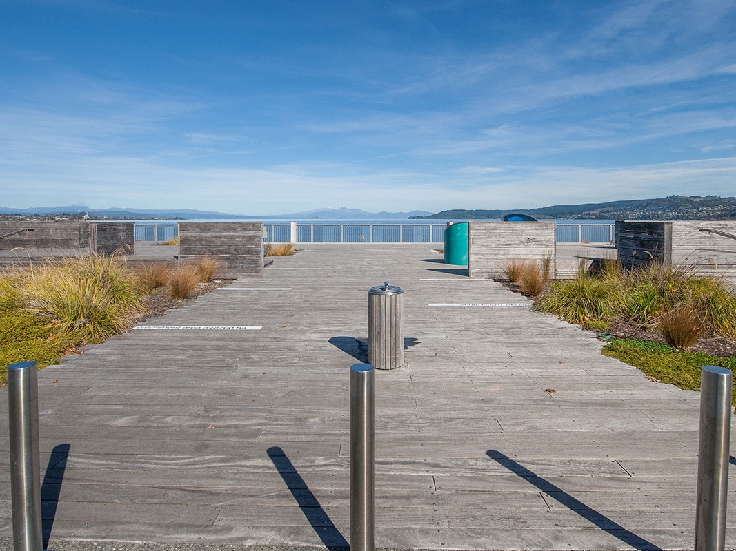 A wooden boardwalk extends towards a waterfront view, framed by landscaped grass and low shrubs. Metal posts and a waste bin are positioned on the deck, offering functional amenities. The distant mountains can be seen across the calm water under a clear blue sky.