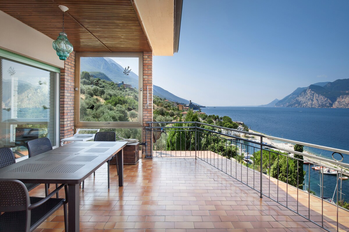 A spacious terrace extends along the apartment, offering stunning views of the lake and surrounding mountains. Outdoor furniture includes a large dining table and chairs, perfect for enjoying meals or relaxing. Bright sunlight enhances the tranquil environment, while the scenic landscape provides a captivating backdrop.