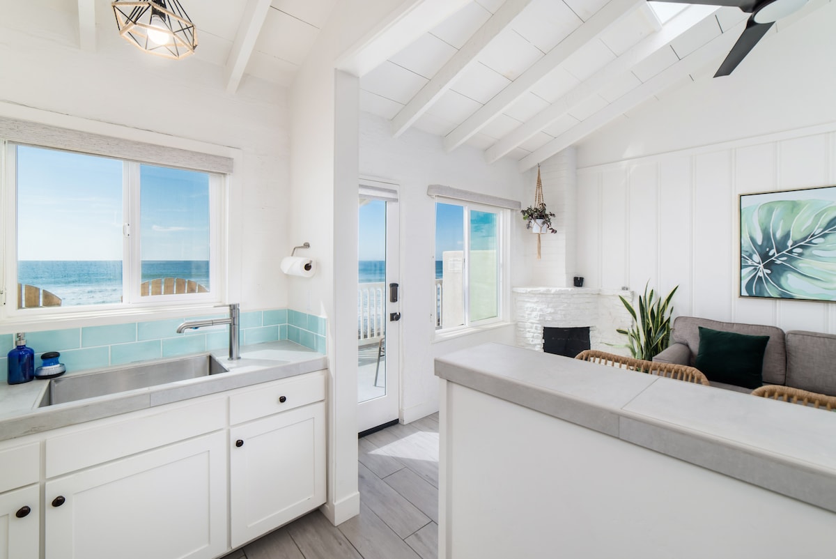 A bright and airy kitchen area features white cabinetry and a large window with ocean views. The adjoining living space includes a comfortable seating arrangement and a decorative plant. Sunlight fills the room, highlighting the open layout and vaulted ceiling.