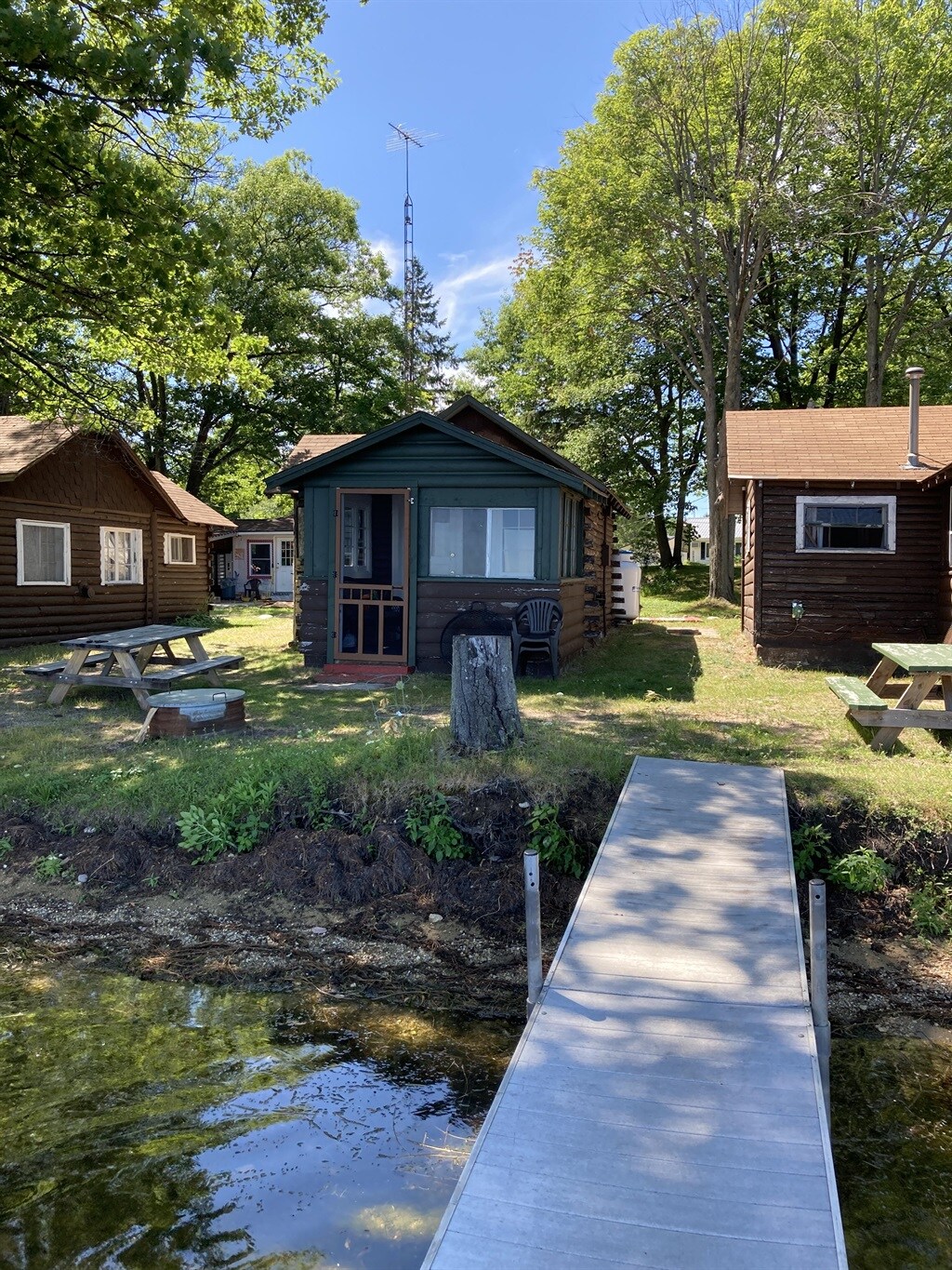 A quaint cabin, nestled among trees, is depicted in the image. The structure features a welcoming porch and is complemented by nearby picnic tables and a small dock extending over the water, providing easy access to the lake. Other rustic cabins are visible in the background.
