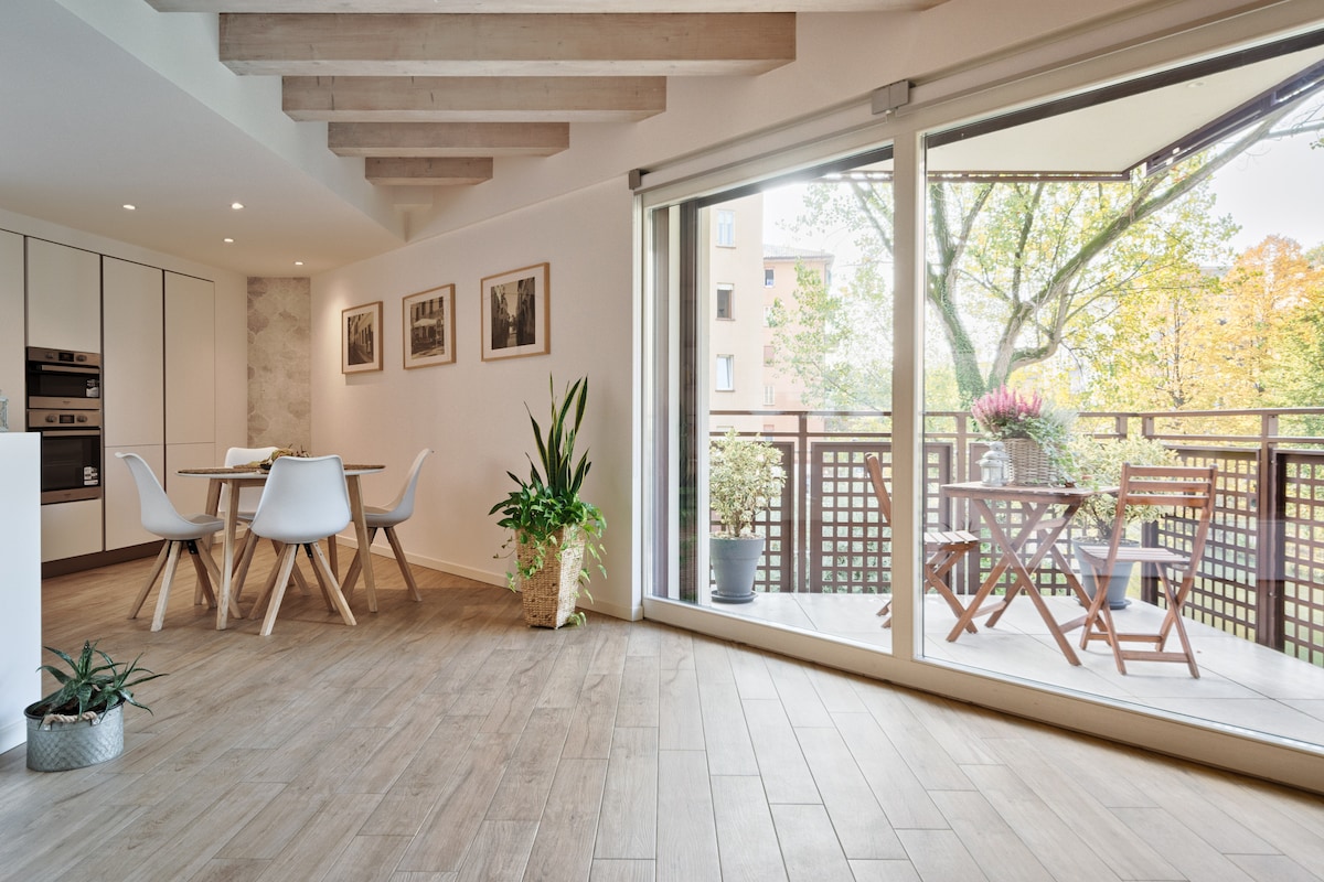 A modern open-plan living area features light wooden beams and tile flooring. A dining table with four chairs is positioned nearby. Large sliding glass doors provide access to a terrace with outdoor seating and potted plants, allowing natural light to fill the room.