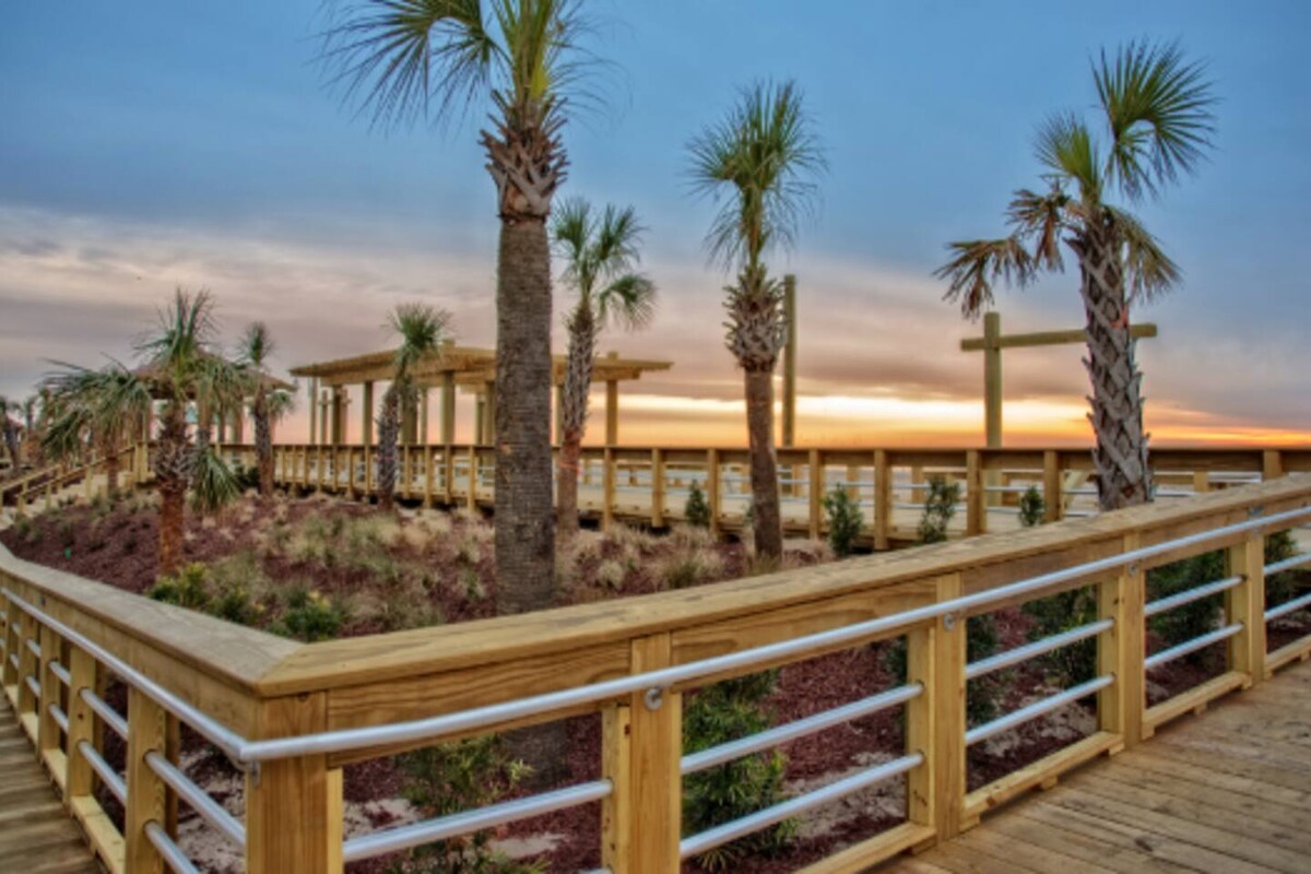 A wooden boardwalk stretches toward a serene horizon, framed by palm trees and lush greenery. The sunset casts soft hues in the sky, while the path leads towards a spacious deck area, enhancing the connection to the natural surroundings.