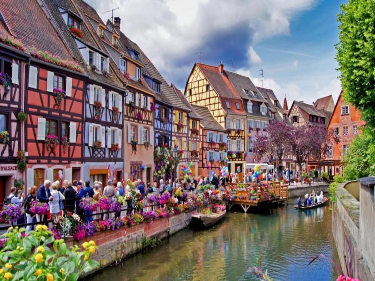 A charming scene along a canal features colorful half-timbered buildings adorned with flower boxes. A group of visitors gathers by a small wooden platform, while boats glide gently on the water. Lush greenery frames the canal adding to the lively atmosphere.
