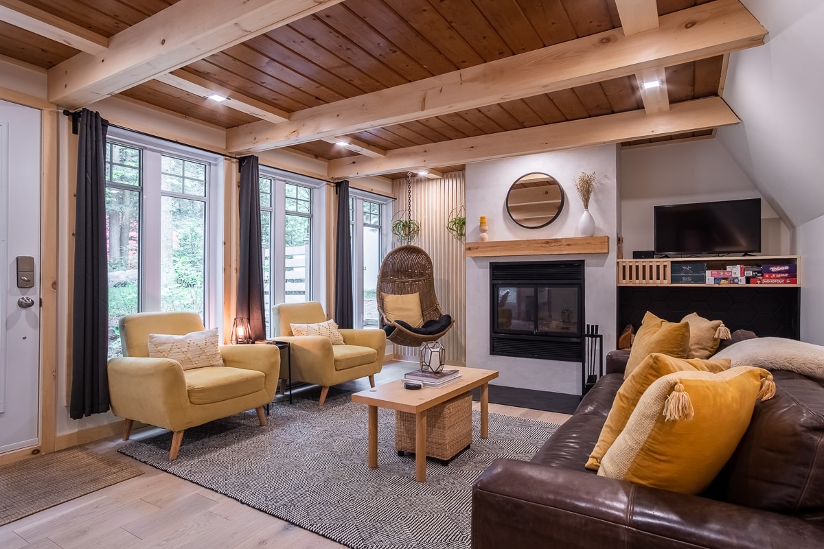 A cozy living area features a mix of textures, with two yellow armchairs and a leather sofa adorned with decorative pillows. A modern fireplace is centered on a wall opposite a television, creating a welcoming space filled with natural light from large windows.