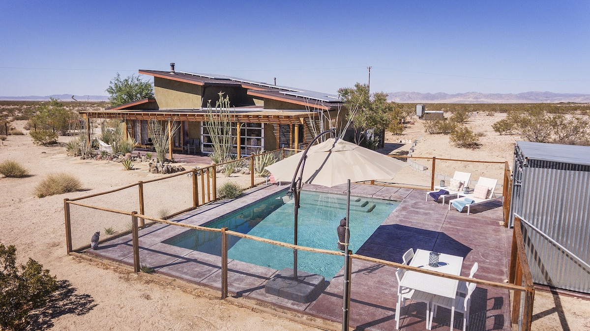 An aerial view captures a contemporary home surrounded by desert landscape. A pool with lounge chairs is situated in the foreground, while a shaded dining area is visible nearby. The structure features large glass windows and surrounding natural vegetation.