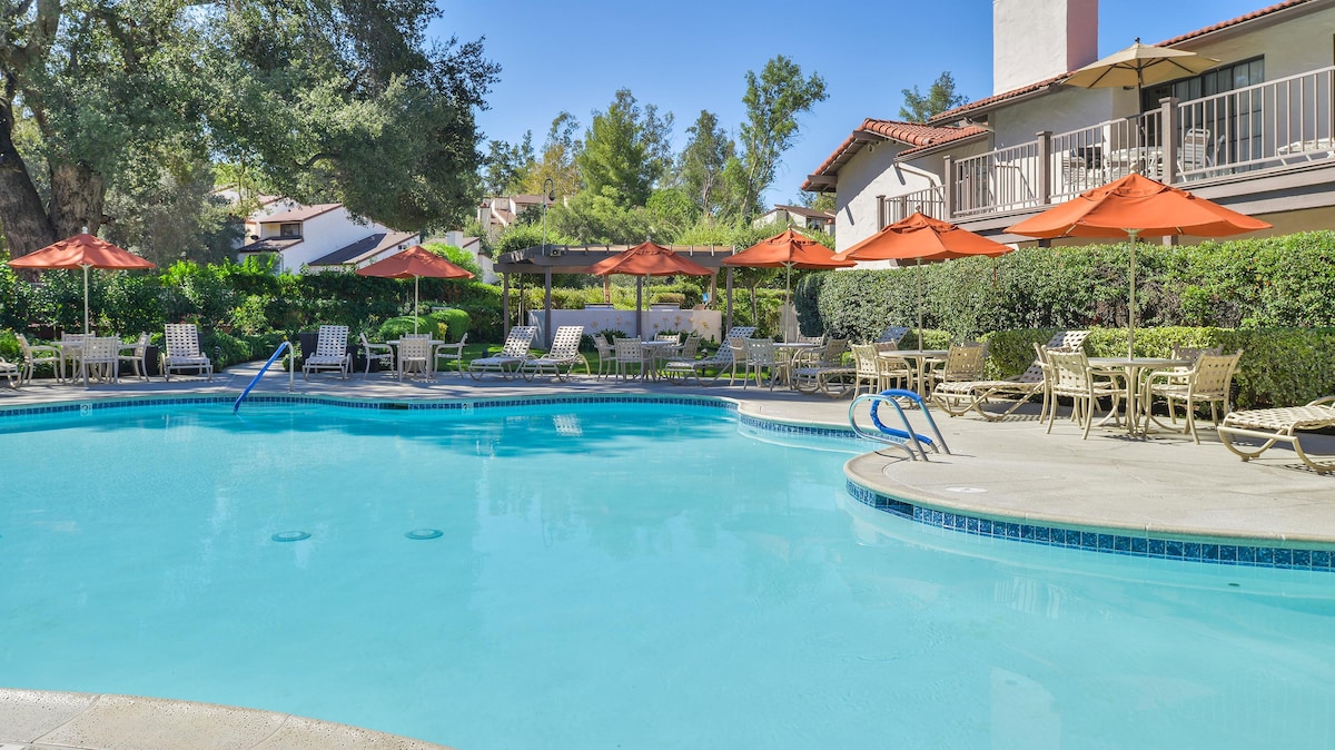 A serene pool area is highlighted by clear blue water surrounded by lounge chairs and umbrellas. The pool area is complemented by well-maintained greenery and the architecture of adjacent buildings, creating a tranquil setting for relaxation.