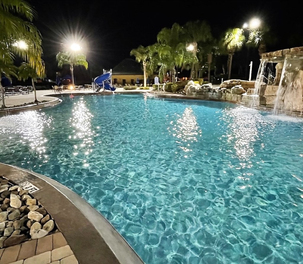 A large, illuminated pool is surrounded by palm trees and lounge chairs during the evening. The water appears clear and inviting, reflecting the bright lights. A water slide and rock features add to the relaxing atmosphere, accompanied by lush landscaping in the background.