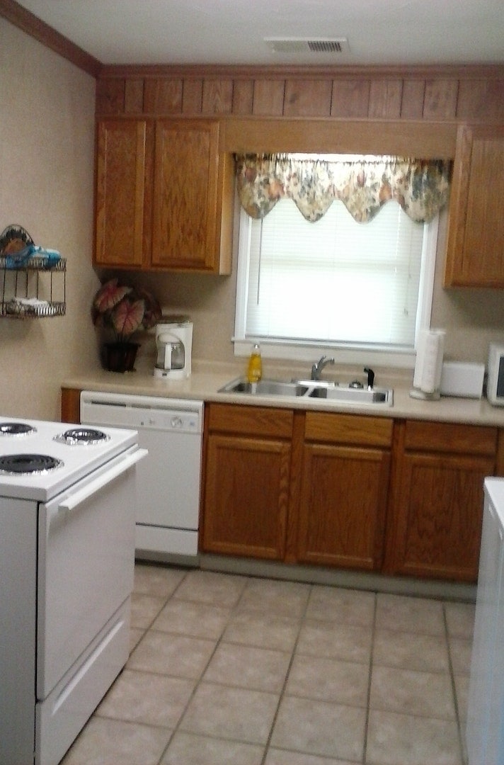 A well-equipped kitchen is designed with wooden cabinetry and a tiled floor. Appliances include a white stove, dishwasher, and microwave. Natural light enters through a window adorned with a floral curtain above the sink, creating a functional and inviting cooking space.