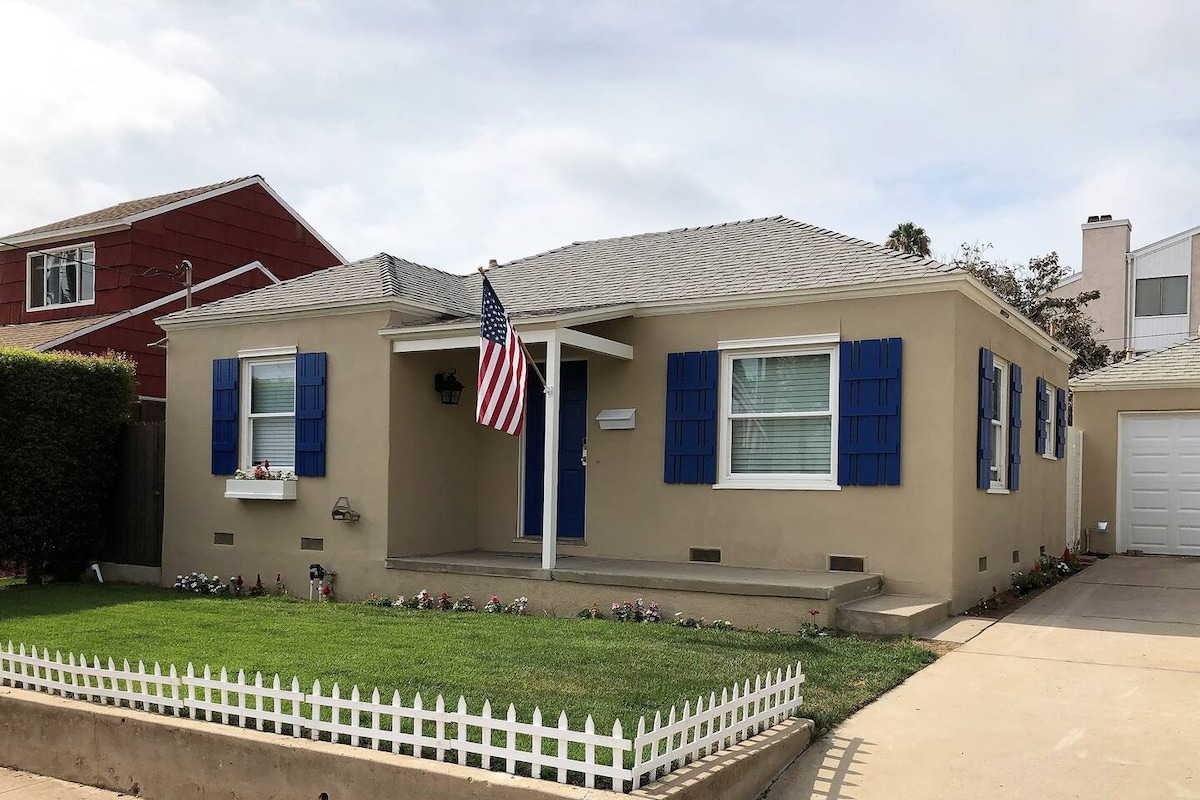 A single-story bungalow features a beige exterior with blue shutters and a front door. An American flag is displayed on the porch, and flowers line the grassy front yard, which is enclosed by a white picket fence. A driveway is visible, leading to a detached garage.