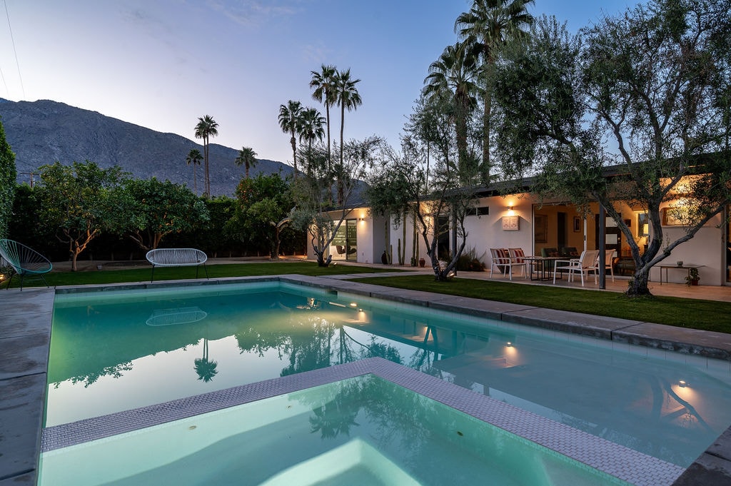 An inviting outdoor space features a clear pool and spa, surrounded by lush landscaping and palm trees. The house is visible in the background with a patio and dining area, framed by mountains and a twilight sky.
