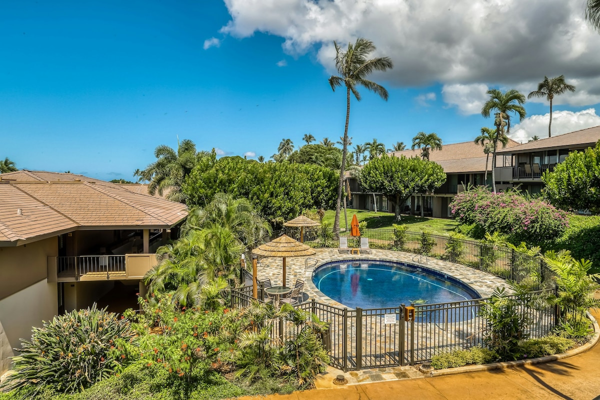 A shared swimming pool is surrounded by lush tropical landscaping and palm trees. Lounge chairs are placed around the pool area, which features a gazebo with a thatched roof. The clear blue sky provides a bright backdrop to the tranquil setting.