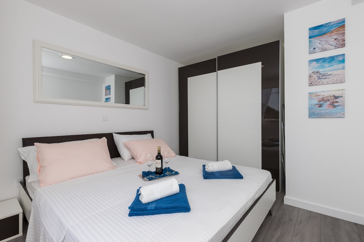 The bedroom features a spacious bed with pale pink accent pillows and neatly arranged towels. A mirror reflects light, enhancing the sense of openness. A wardrobe provides storage, while colorful coastal artwork adds visual interest to the walls.