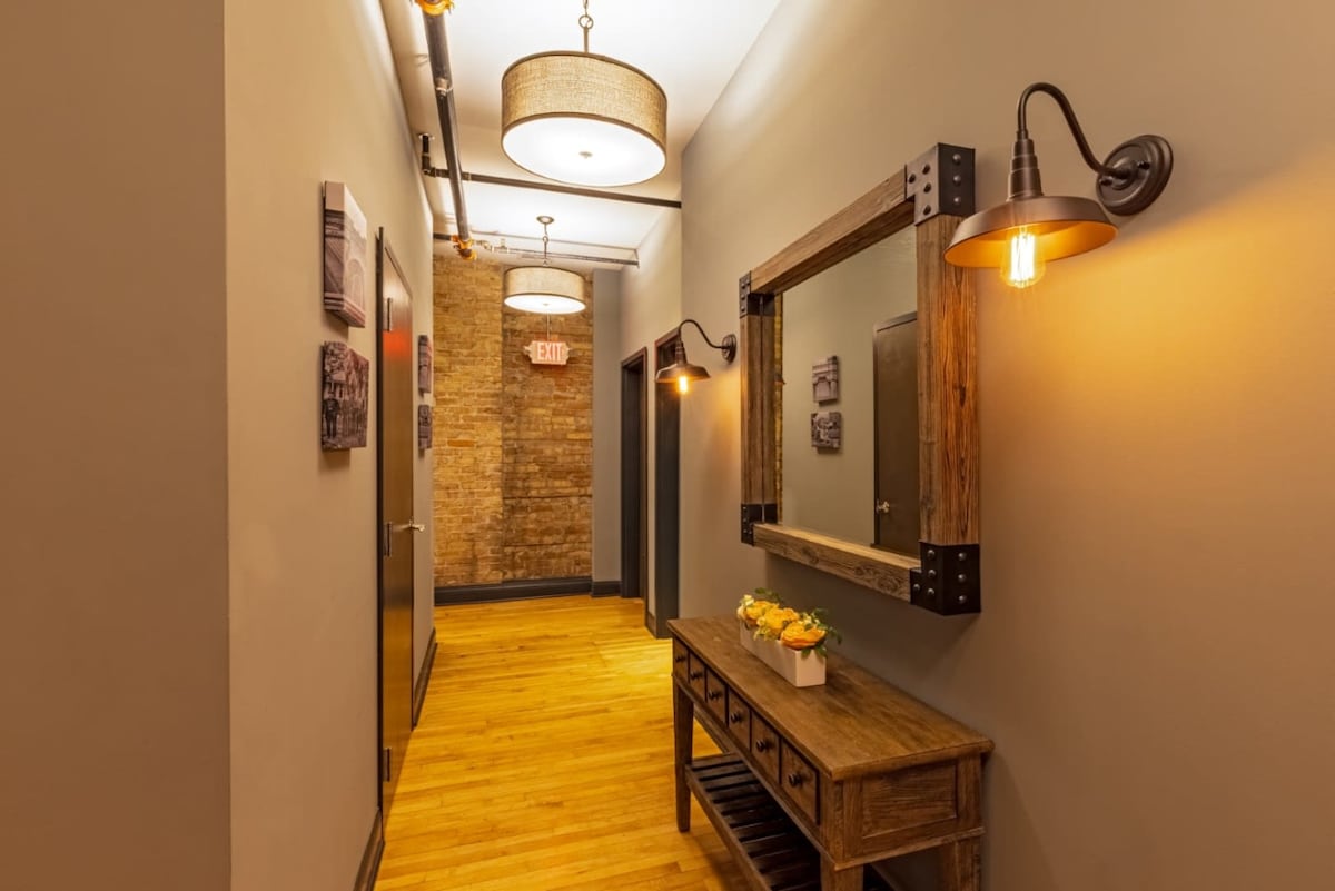 An inviting hallway features exposed brick walls and wooden flooring. Soft lighting is provided by stylish ceiling fixtures and wall-mounted sconces. A rustic wooden console table holds a vase of flowers, complementing the warm neutral tones throughout the space.