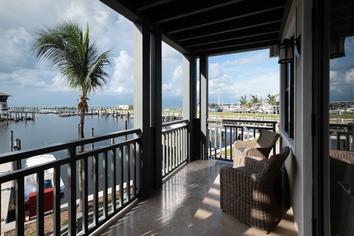A private terrace offers views of the marina, framed by a tall palm tree. Two woven chairs provide a cozy seating area, while the water and boat slips are visible in the background under a partly cloudy sky.