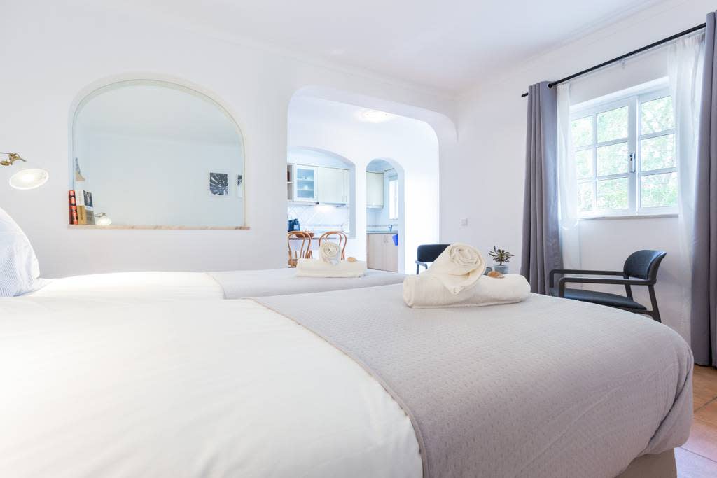 The studio apartment features two single beds positioned side by side, accompanied by neatly rolled towels. An archway leads to a fully equipped kitchen, while natural light filters through the nearby window, enhancing the open and airy environment.