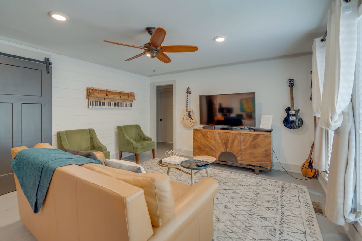 A welcoming living area features a beige sofa complemented by two green armchairs. A wooden console table stands against the wall, hosting a large flat-screen TV. A ceiling fan provides airflow, while natural light filters through sheer curtains draped by the windows.