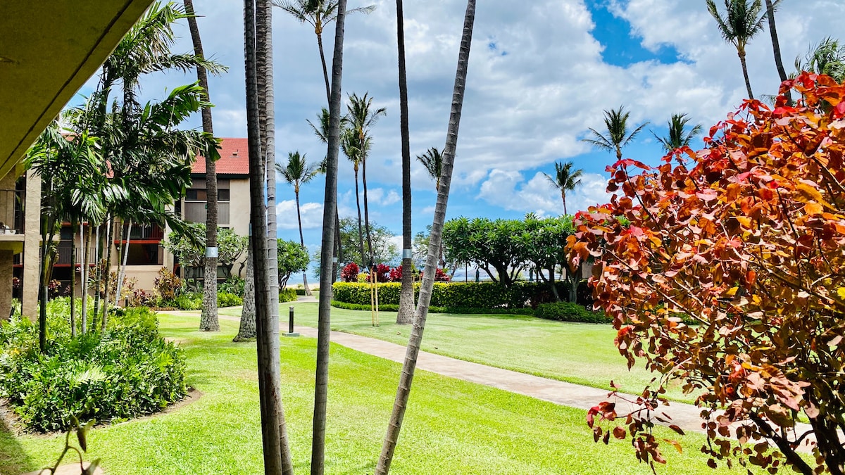 A lush garden area is depicted with tropical plants and palm trees swaying gently in the breeze. Green grass stretches towards the horizon, where a glimpse of the ocean can be seen through the foliage. Bright flowers add splashes of color to the scenery.