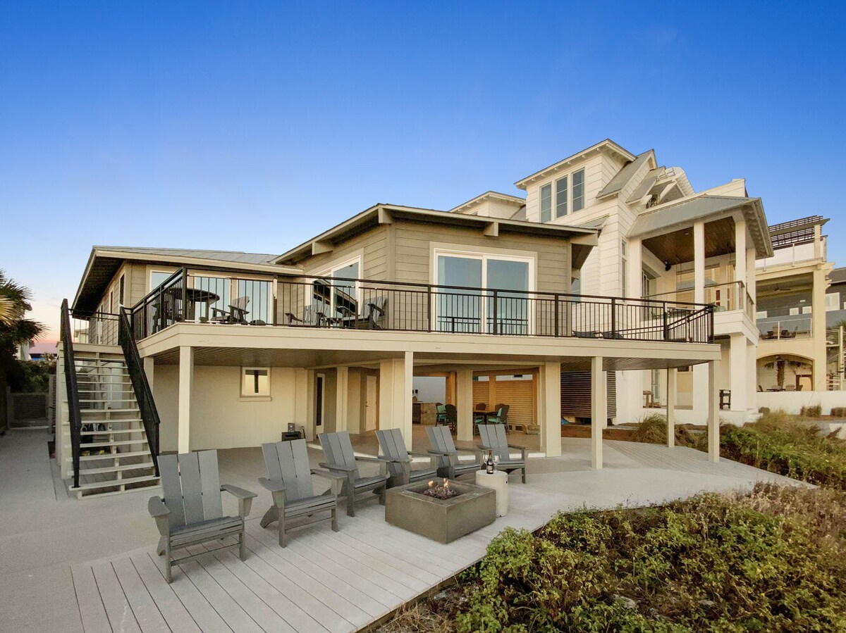 The exterior of the beachfront home is presented, featuring a spacious deck with multiple lounge chairs and a fire pit. The multi-story structure showcases large windows and ample outdoor seating, creating inviting spaces for relaxation. Lush coastal vegetation is visible in the foreground.