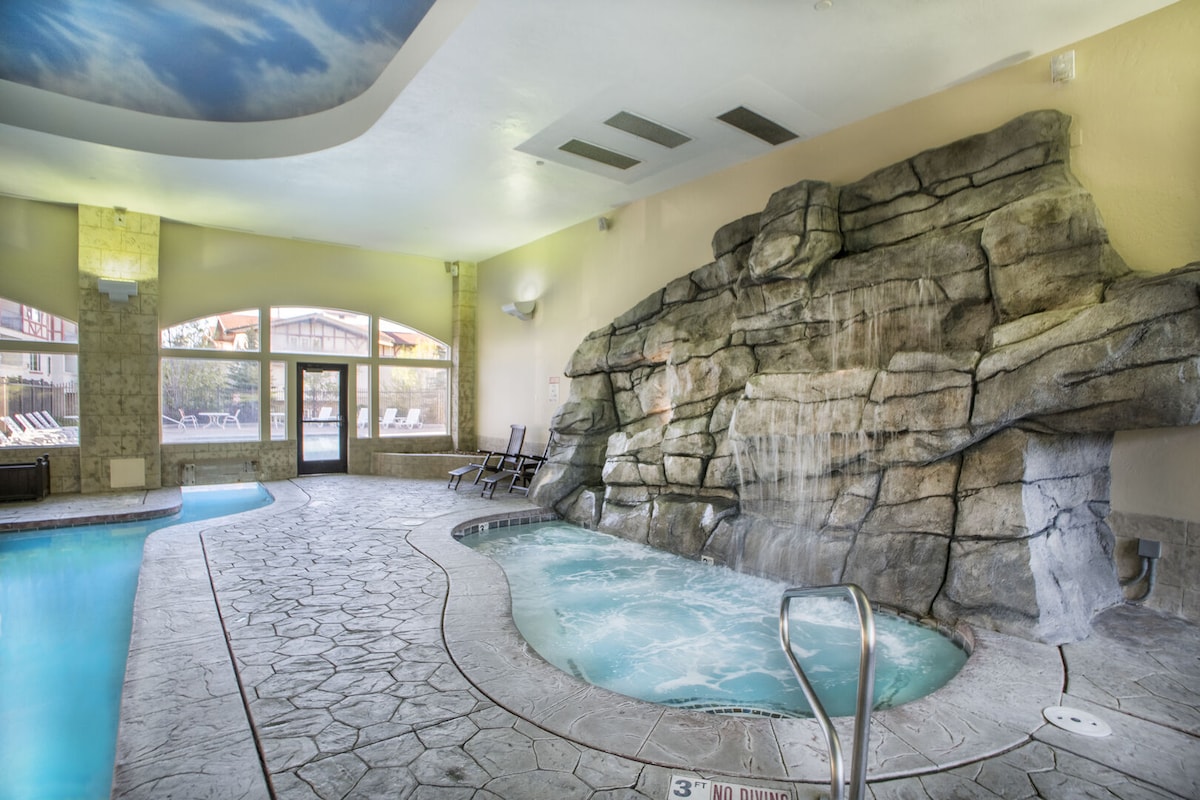 An indoor pool area features a natural rock waterfall and a spacious hot tub, surrounded by textured stone walls. Large windows allow natural light to fill the space, showcasing lounge chairs nearby and a serene atmosphere for relaxation.