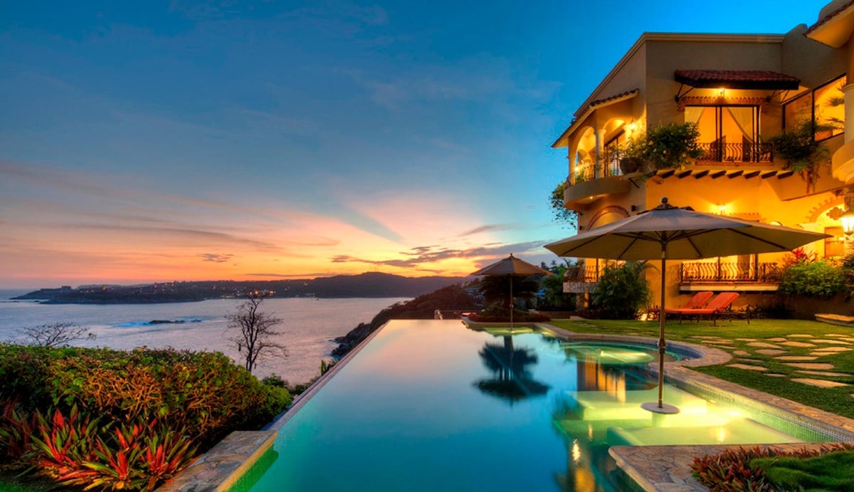 An oceanfront infinity pool is seen at dusk, blending with the horizon. Umbrellas provide shade over lounge chairs, while lush tropical gardens frame the space. The exterior of a multi-level villa can be viewed, illuminated by soft lighting, against a vibrant sunset sky.