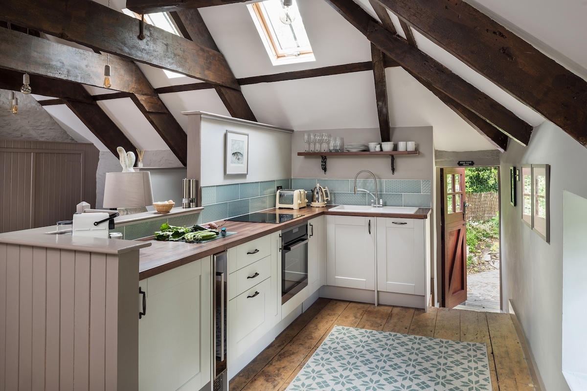 An open-plan kitchen area features vaulted ceilings with exposed wooden beams and a wooden floor. Equipped with modern appliances, including an oven and sink, the space is complemented by a centrally placed wooden island and skylights allowing natural light to fill the room.