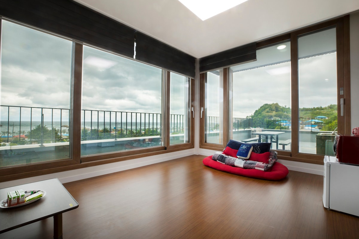 A spacious living area features large floor-to-ceiling windows that offer expansive views of the surrounding landscape. A red cushion is laid against the wall, providing a cozy seating option. Natural light brightens the room, highlighting the wooden flooring.