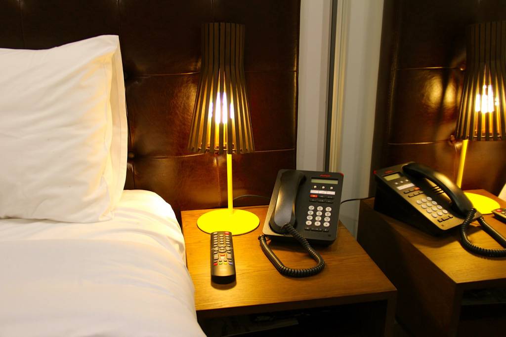 A bedside table is shown with two telephones and a remote control. A stylish lamp with a warm glow complements the soft texture of the headboard. Crisp white bedding is visible, enhancing the clean and modern aesthetic of the space.
