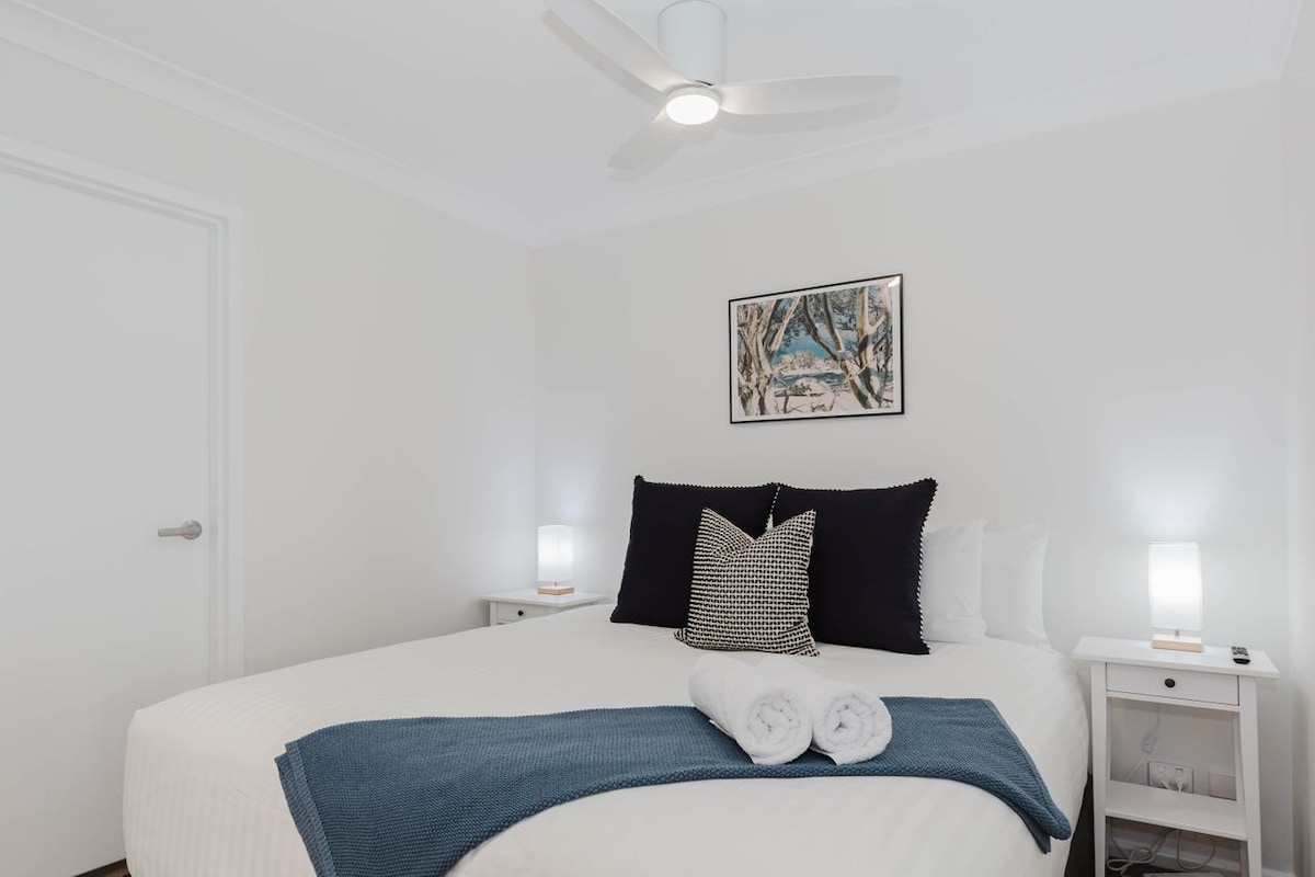 A well-lit bedroom features a spacious bed with crisp white linens and a textured navy throw draped across. Two bedside tables with lamps are positioned on either side of the bed. An artwork is displayed above the bed, adding a touch of decor to the neutral walls.