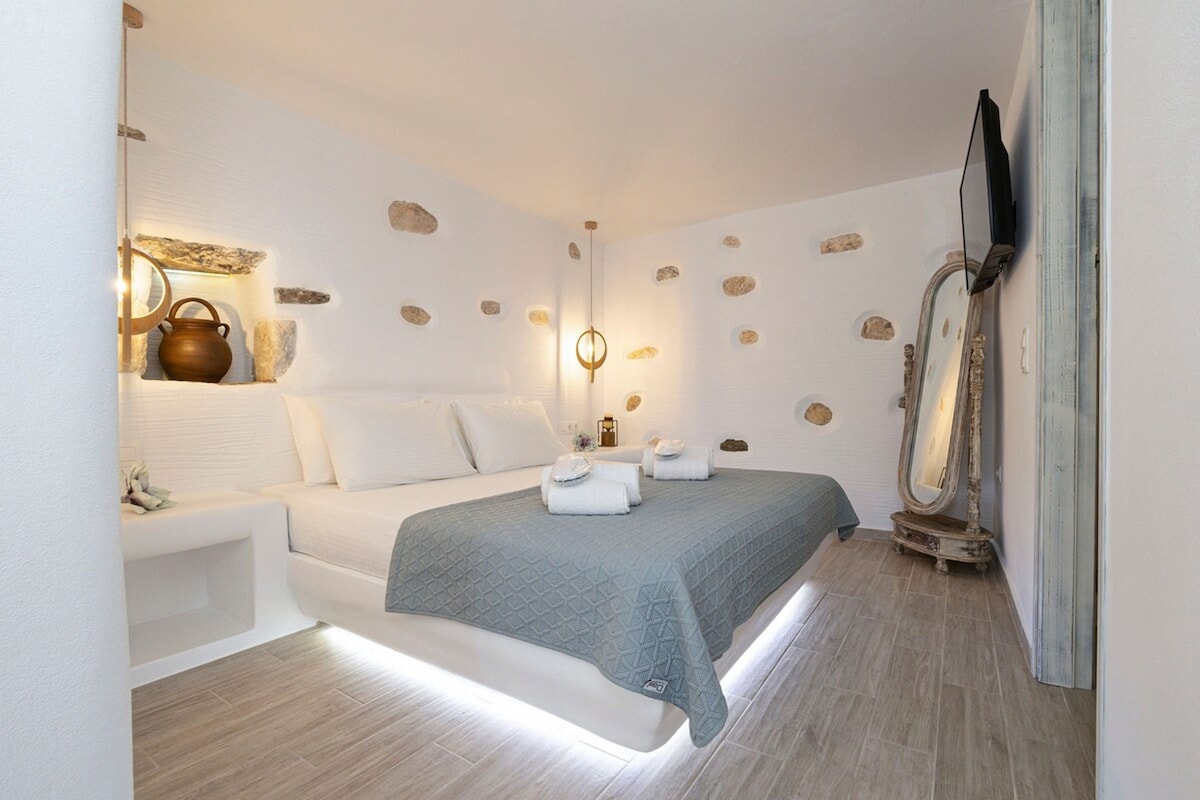 The master bedroom features a spacious bed with a light quilt and rolled towels. Soft lighting illuminates the area, highlighting the stone details on the walls. A full-length mirror and a mounted television are present, complementing the serene atmosphere.