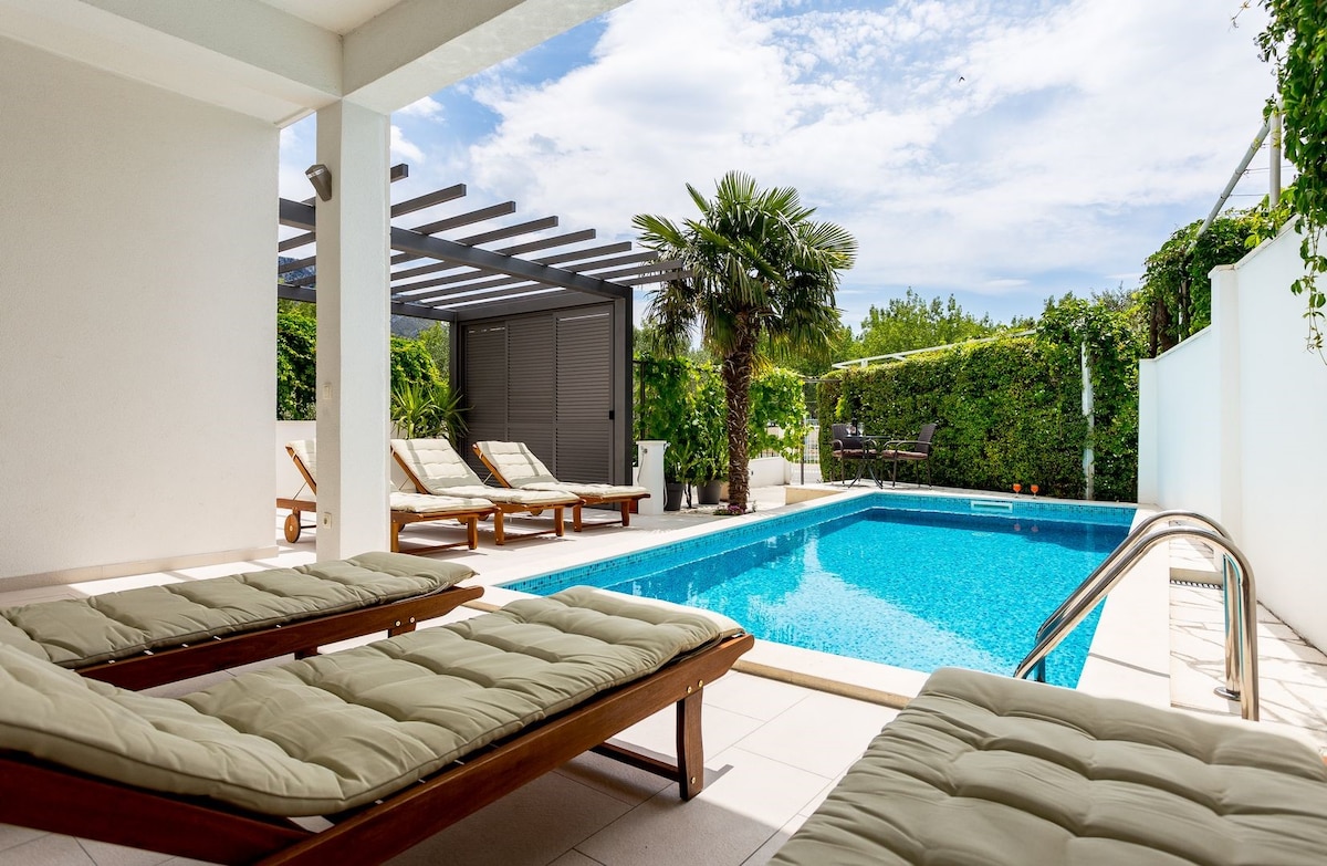 A private pool area features a rectangular pool surrounded by a patio. Sun loungers with soft cushions are positioned beside the pool, offering space for relaxation. Lush greenery and palm trees provide shade, enhancing privacy in this outdoor oasis.