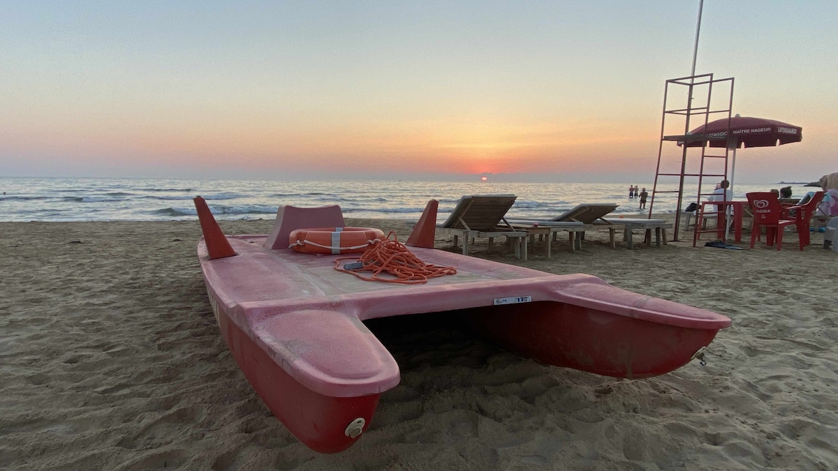 A vibrant red catamaran rests on sandy beach, surrounded by gentle waves and a colorful sunset in the background. Beach chairs and umbrellas can be seen in the distance, hinting at a relaxed coastal atmosphere.