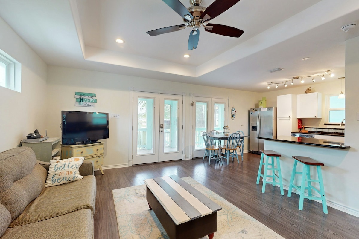 The living area features a comfortable sofa facing a flat-screen TV, with natural light entering through multiple glass doors. A dining table for four and a kitchen island with seating for two are visible. Coastal decor is present in the space.