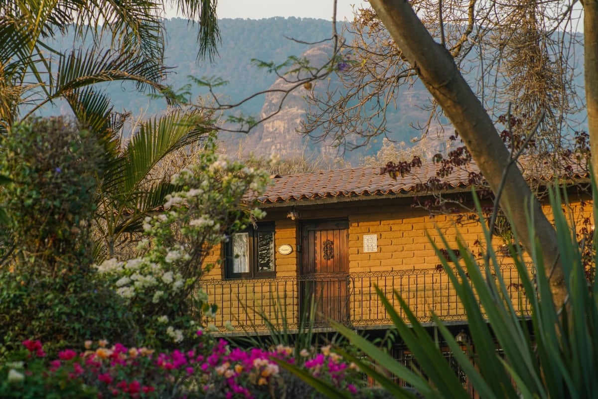 A rustic cabin is nestled among vibrant gardens filled with flowers. Lush greenery surrounds the entrance, featuring a wooden door that complements the natural tones of the cabin. The backdrop showcases a mountainous landscape, creating a serene and inviting outdoor atmosphere.