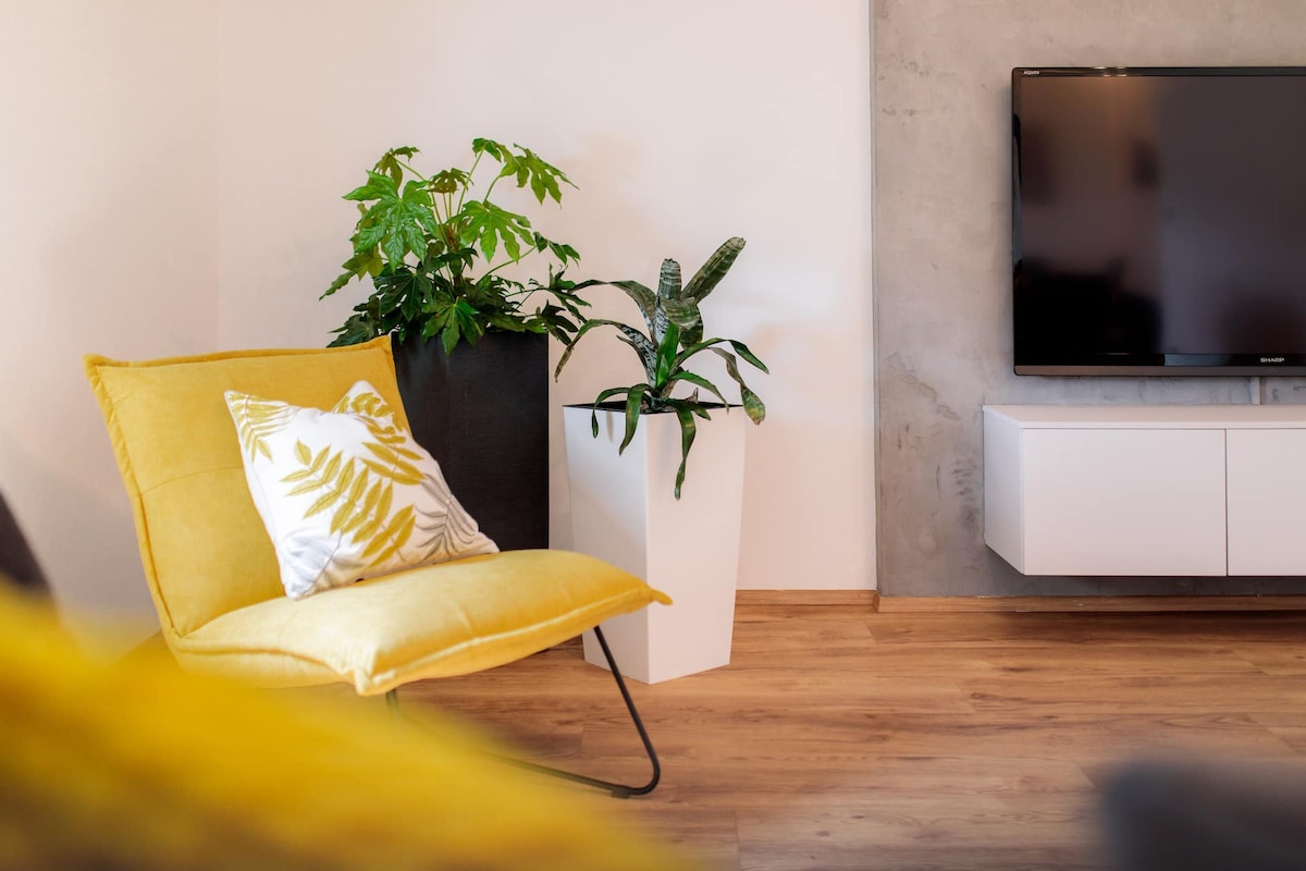 A vibrant yellow chair is positioned next to a sleek white television cabinet, with a flat-screen TV mounted on the wall. Potted plants in contrasting planters add a touch of greenery to the warm wooden flooring and soft, neutral walls.