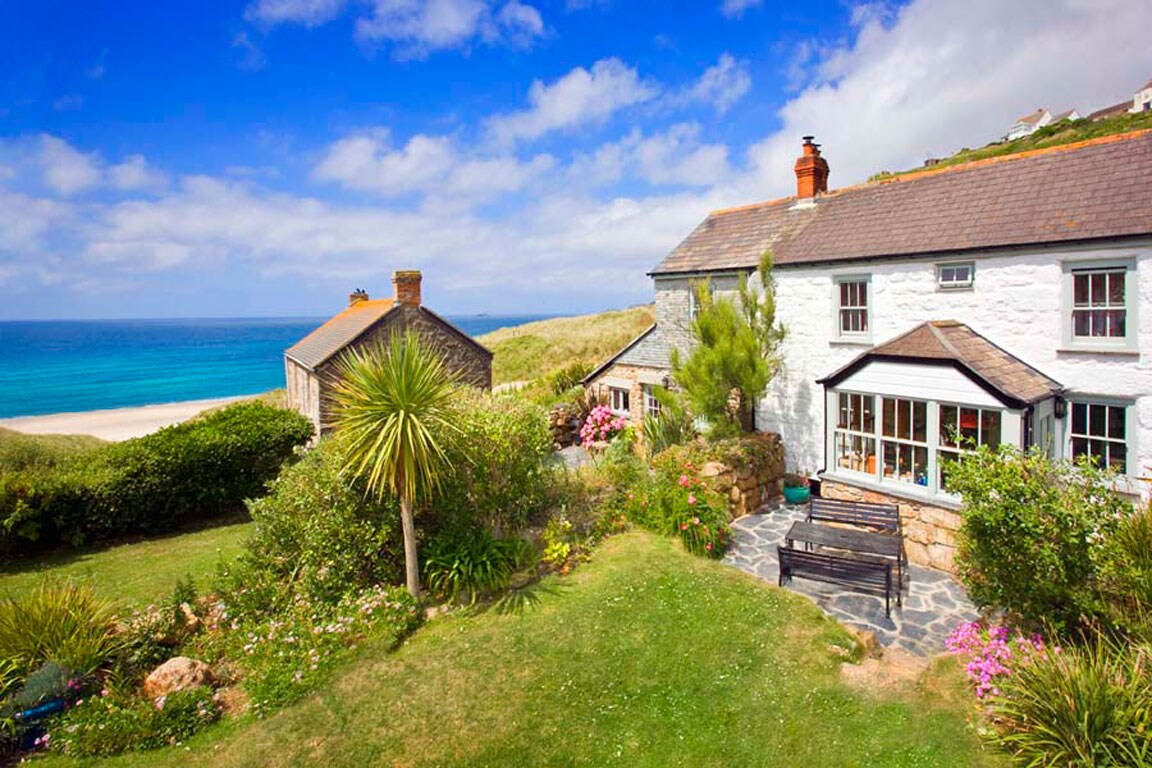 A traditional Cornish cottage is seen nestled in a lush garden, featuring vibrant blooms and manicured greenery. A nearby bench offers a picturesque view of the sandy beach and crystal blue sea, under a sky with wispy clouds, creating a serene coastal atmosphere.