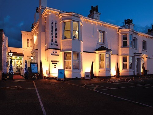 A charming Grade II listed hotel is displayed at dusk, illuminated by warm lights that highlight the architectural details. The exterior features a combination of white walls and decorative trim, flanked by neatly arranged conical trees along the entrance.