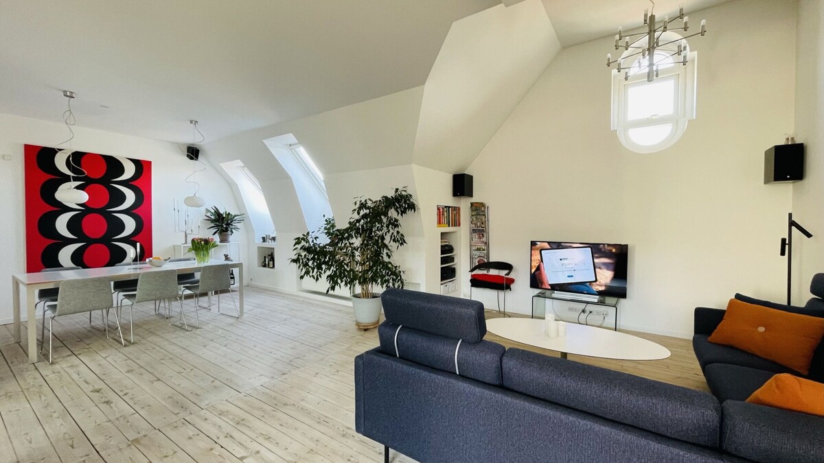 The open-plan living space features a comfortable sofa facing a television and a dining area with a table and chairs. Large, sloped windows allow plenty of natural light, while a potted plant adds a touch of greenery to the room.