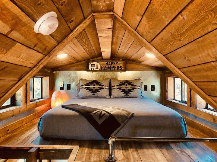 Tucked under the wood-beamed ceiling, the loft bedroom features a plush king bed, southwestern-patterned pillows, and a playful "Happy Campers" sign overhead — cozy, characterful, and made for a good night's sleep.
