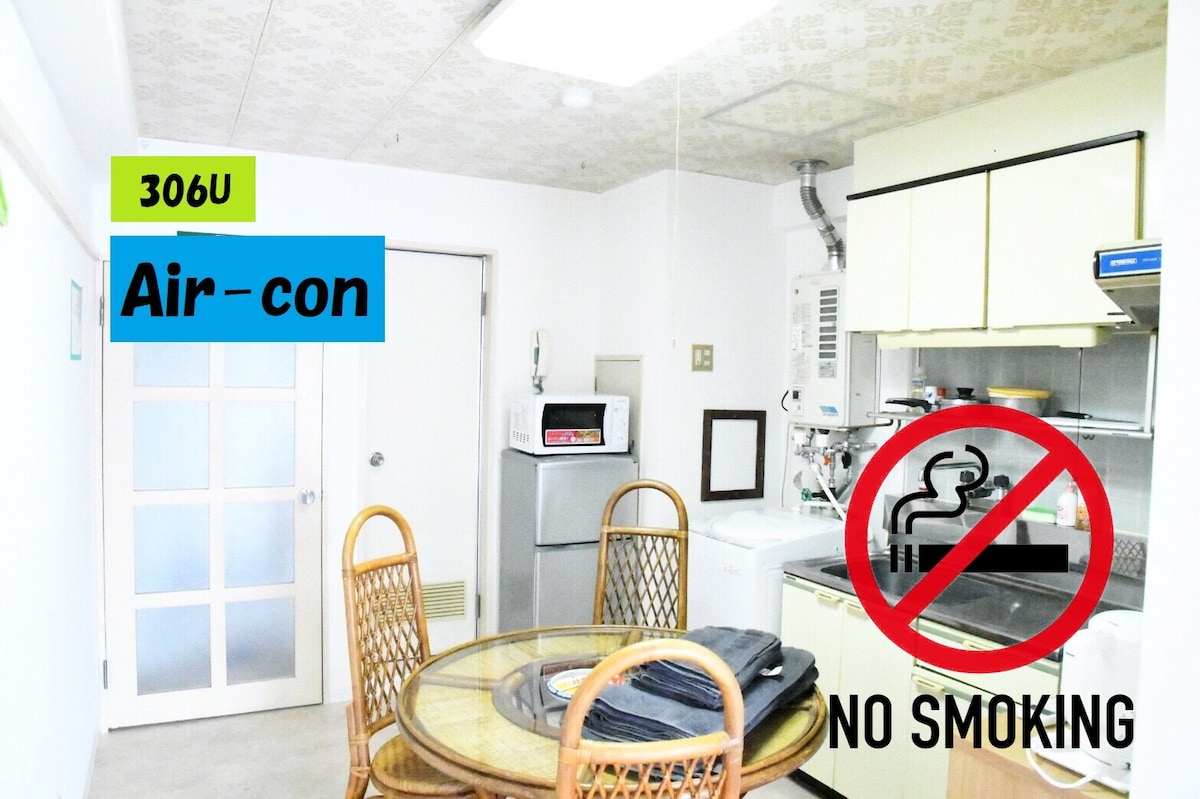 A bright kitchen area is visible, featuring a round table surrounded by rattan chairs. The space includes a microwave, sink, and cooking appliances, with light-colored cabinets above. A no smoking sign is prominently displayed, and air conditioning is indicated in the room.