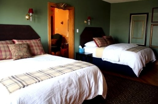 A cozy room features two Queen beds with decorative headboards and plaid accents. The walls are painted a calming green, complemented by warm lighting from wall sconces. A doorway leads to a sitting area, showcasing additional furnishings in the background.