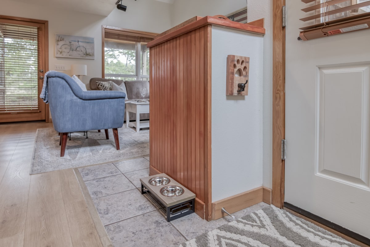 A welcoming entry area showcases a wooden console with a dog feeding station featuring two stainless steel bowls. Natural light enters from a nearby window, illuminating the textured rug and the comfortable seating area visible in the background.