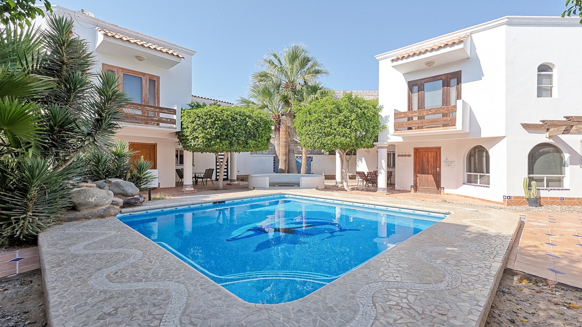 A clear blue pool surrounded by landscaped greenery is featured in the center. Two balconies overlook the pool area, providing a glimpse of the outdoor seating arrangement. The property is framed by palms and shrubs, contributing to the serene atmosphere.